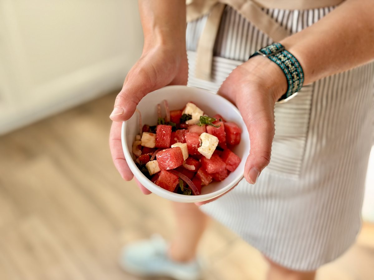 Wider view of a woman in a striped apron cradling a white bowl of watermelon and feta salad, her legs and the wood floor visible b