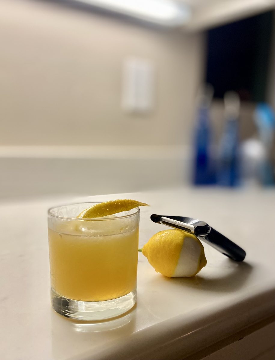 A cloudy pale-yellow cocktail in a short rocks glass with a lemon twist, beside a partly peeled lemon and a channel knife on a whi