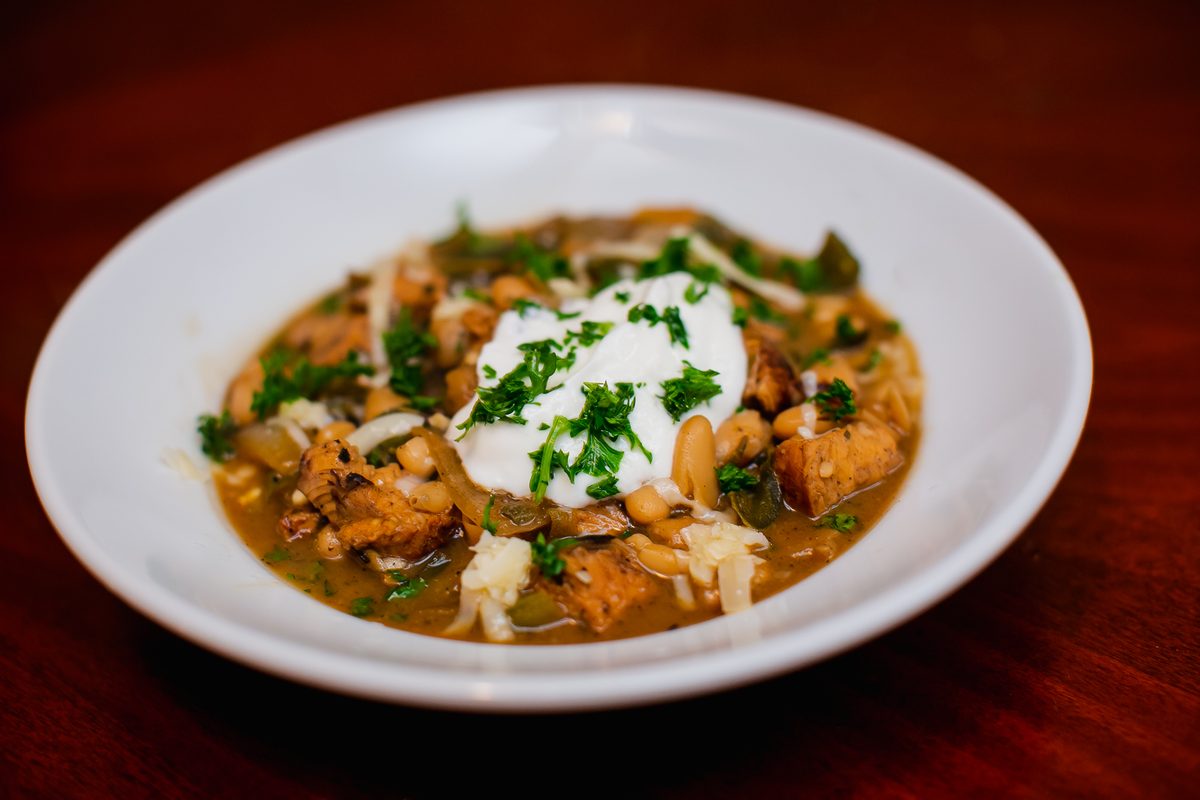 A wide white bowl of white bean chicken chili with visible chunks of chicken, shredded cheese, sour cream and chopped cilantro, on