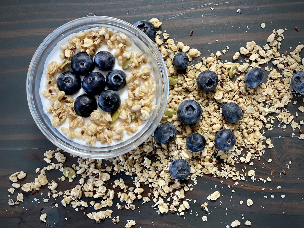 Overhead shot of a small glass bowl of yogurt topped with oat granola and fresh blueberries, with more granola and berries scatter