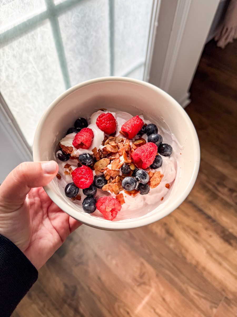 Two hands hold a white bowl of pink-tinted yogurt topped with granola, raspberries and blueberries in front of a bright window wit