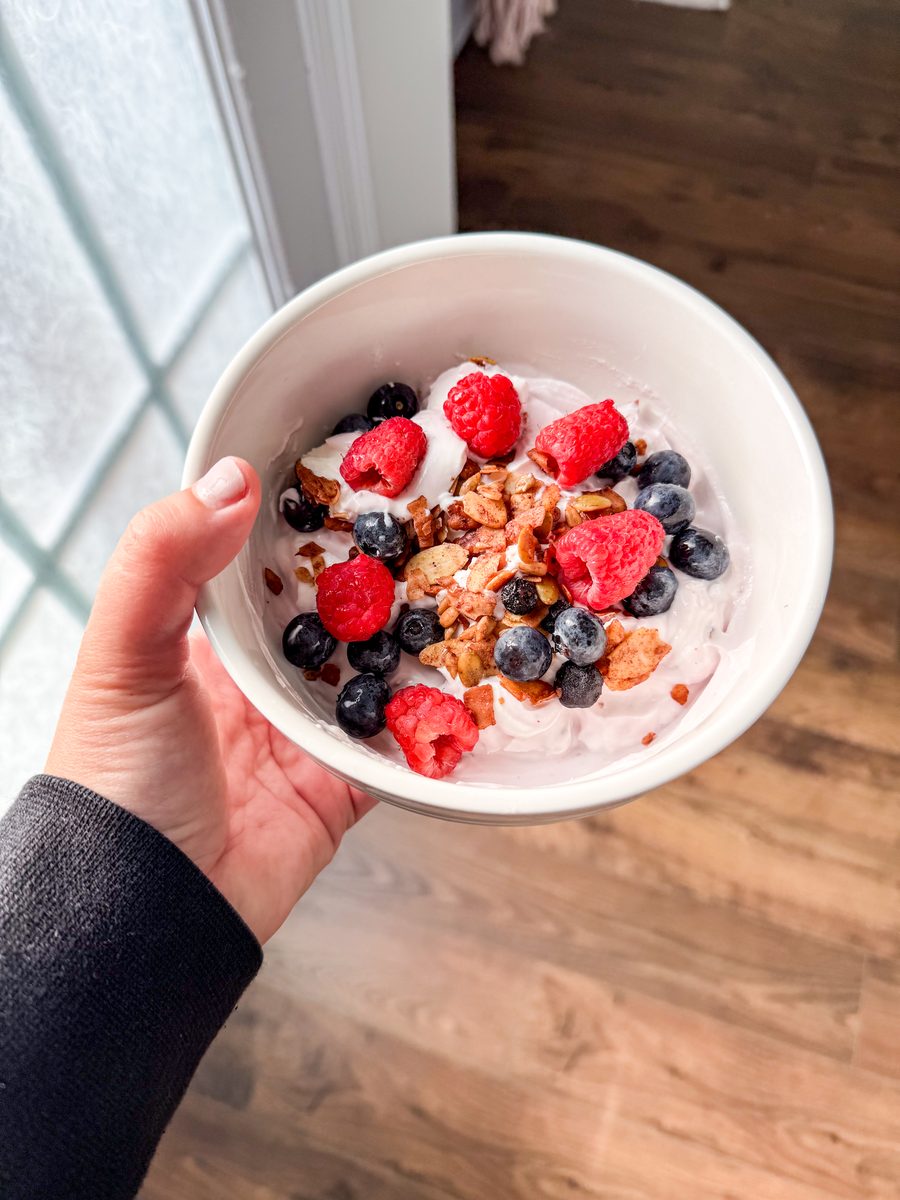 A hand in a dark sleeve holds a white bowl of yogurt with granola, raspberries and blueberries beside a window, wood floor and doo