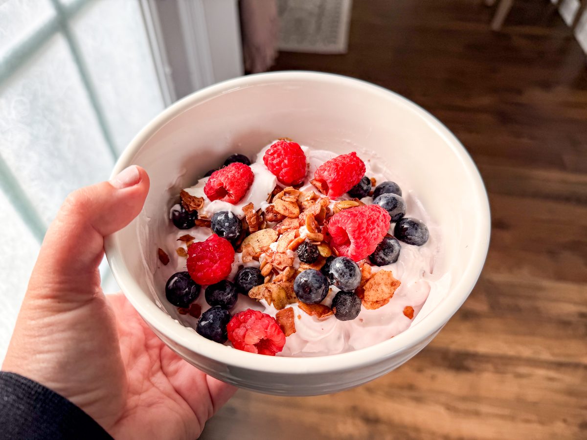 Close-up of a hand holding a white bowl of pink yogurt topped with granola, raspberries and blueberries, window light and wood flo