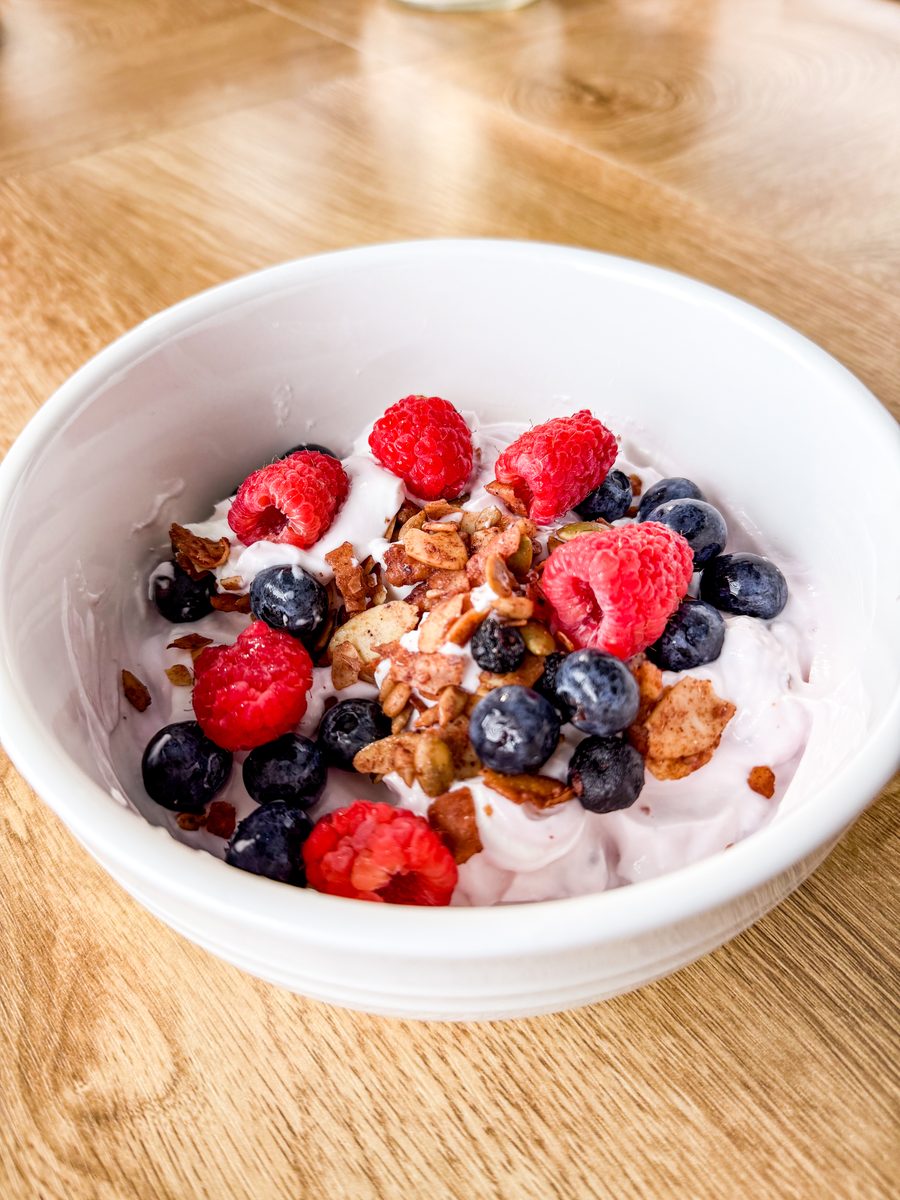 A white bowl of yogurt topped with granola, raspberries and blueberries sitting on a light wood table, shot from a high angle.