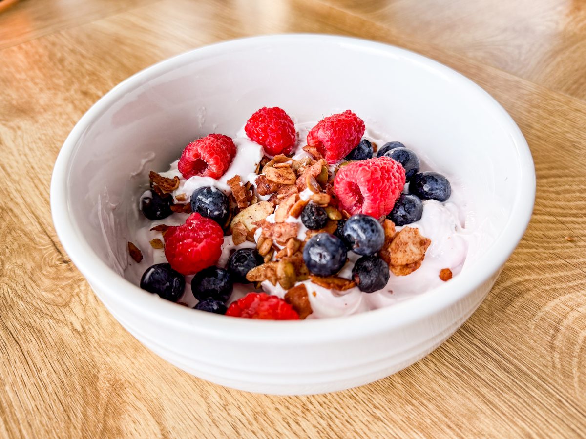 Close overhead view of a white bowl of yogurt heaped with granola clusters, raspberries and blueberries on a light oak table.