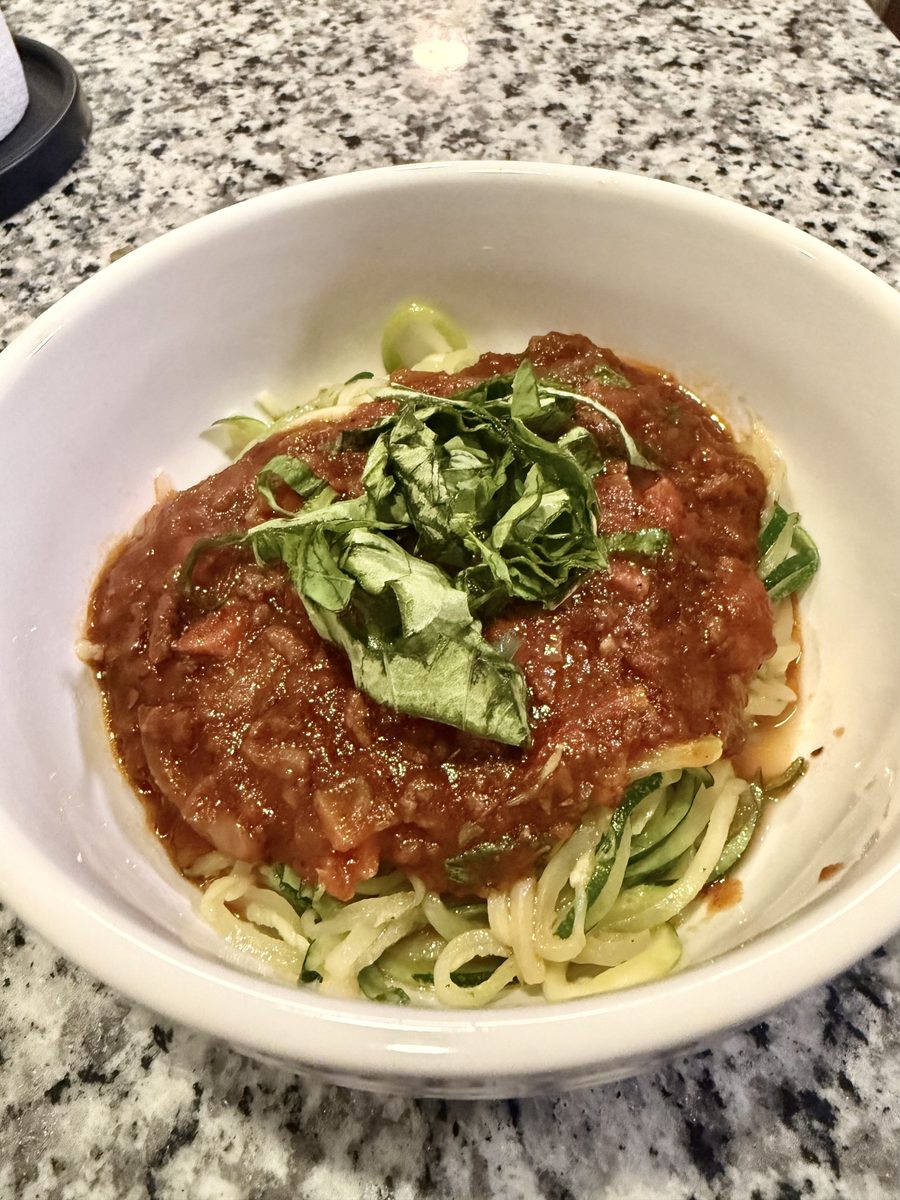 A white bowl of zucchini noodles topped with a thick tomato sauce and shredded basil on a speckled granite countertop; the sauce i