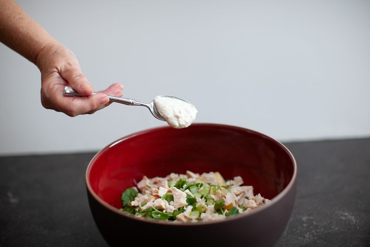 A hand holds a spoon of mayonnaise above a dark red bowl of diced chicken salad with celery and herbs on a dark counter against a 