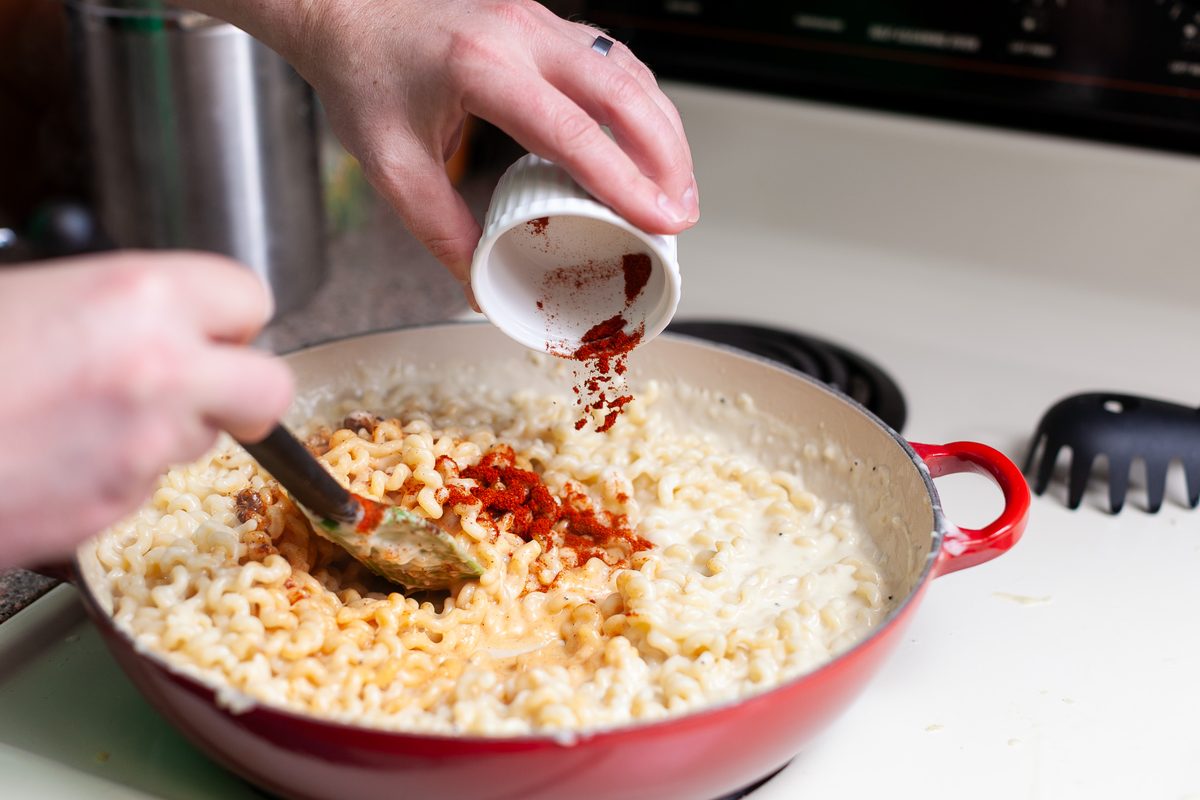 Close view of two hands pouring paprika from a white ramekin into a red pan of creamy macaroni and cheese on a stovetop, spatula i