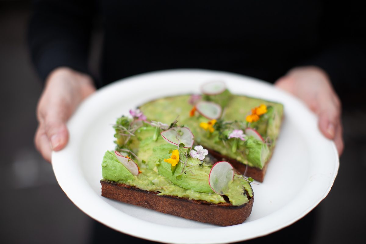 Hands hold a white plate with two slices of dark rye avocado toast topped with smashed avocado, radish slices, edible flowers and