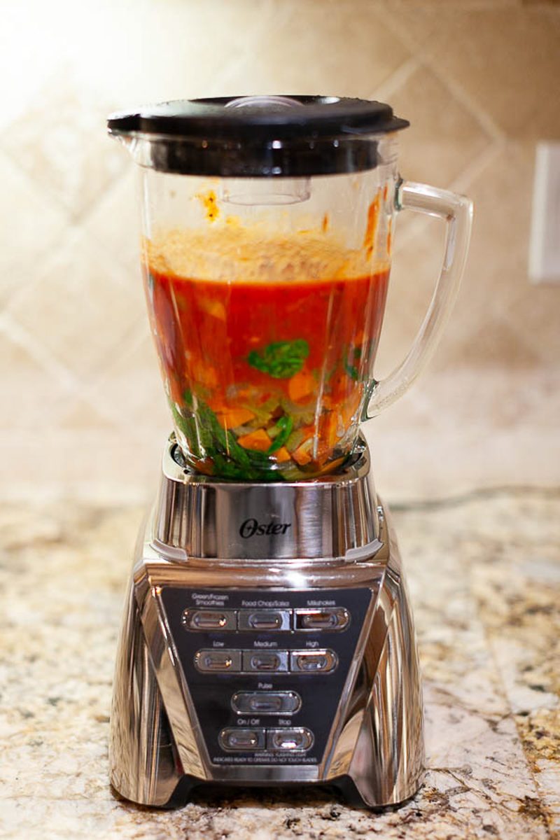 A chrome Oster blender on a granite counter, its jar filled with red-orange tomato and carrot soup with cilantro leaves not yet bl