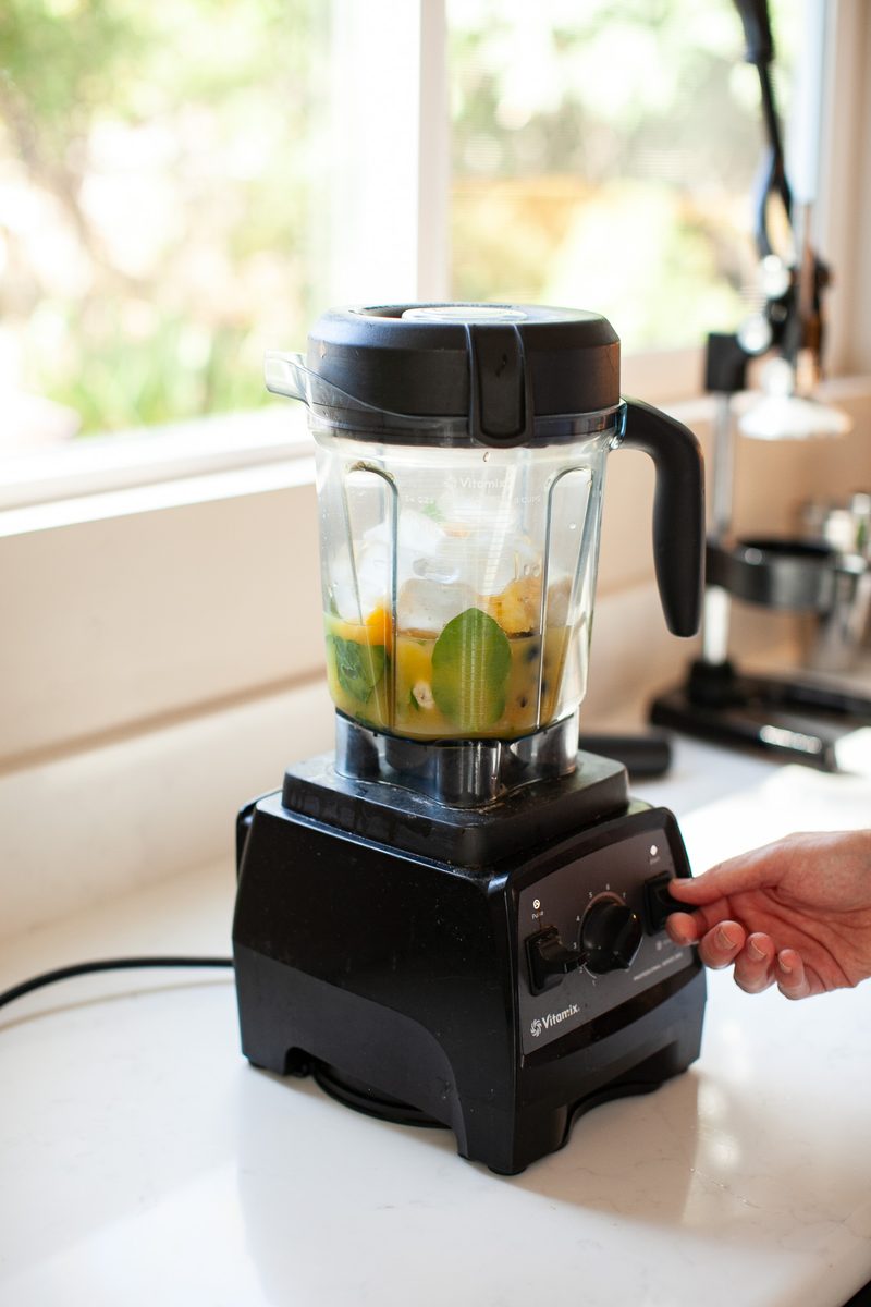 A hand reaches for the controls of a black blender by a sunlit window, the jar holding citrus and green leaves for a smoothie.