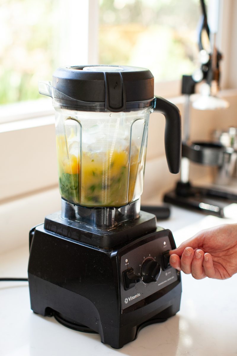 A blender on a white counter in front of a bright window, contents part blended into a yellow-green mixture, a hand at the base co