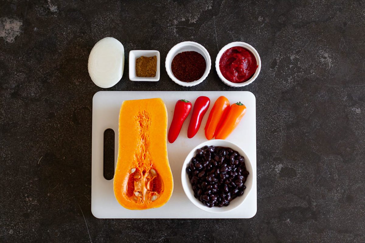 Overhead flat lay of chili ingredients on a dark counter: halved butternut squash, mini sweet peppers and black beans on a white c
