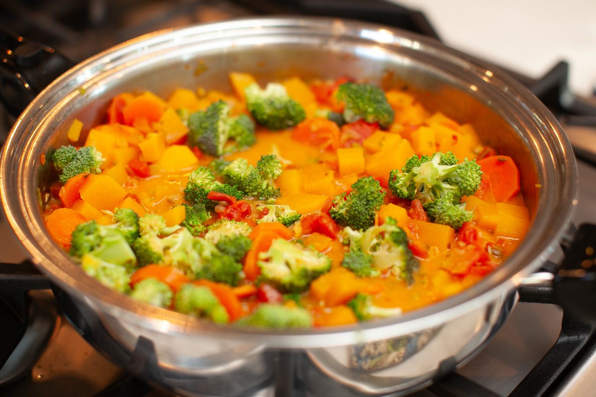 A stainless steel skillet on a gas stove filled with a finished curry of butternut squash cubes, broccoli florets and red pepper i