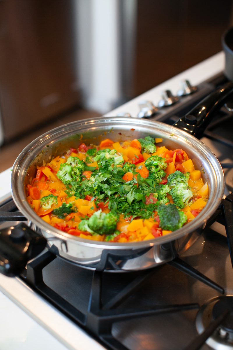 A stainless skillet of butternut squash and broccoli curry topped with chopped cilantro, sitting on a gas cooktop with a dim kitch