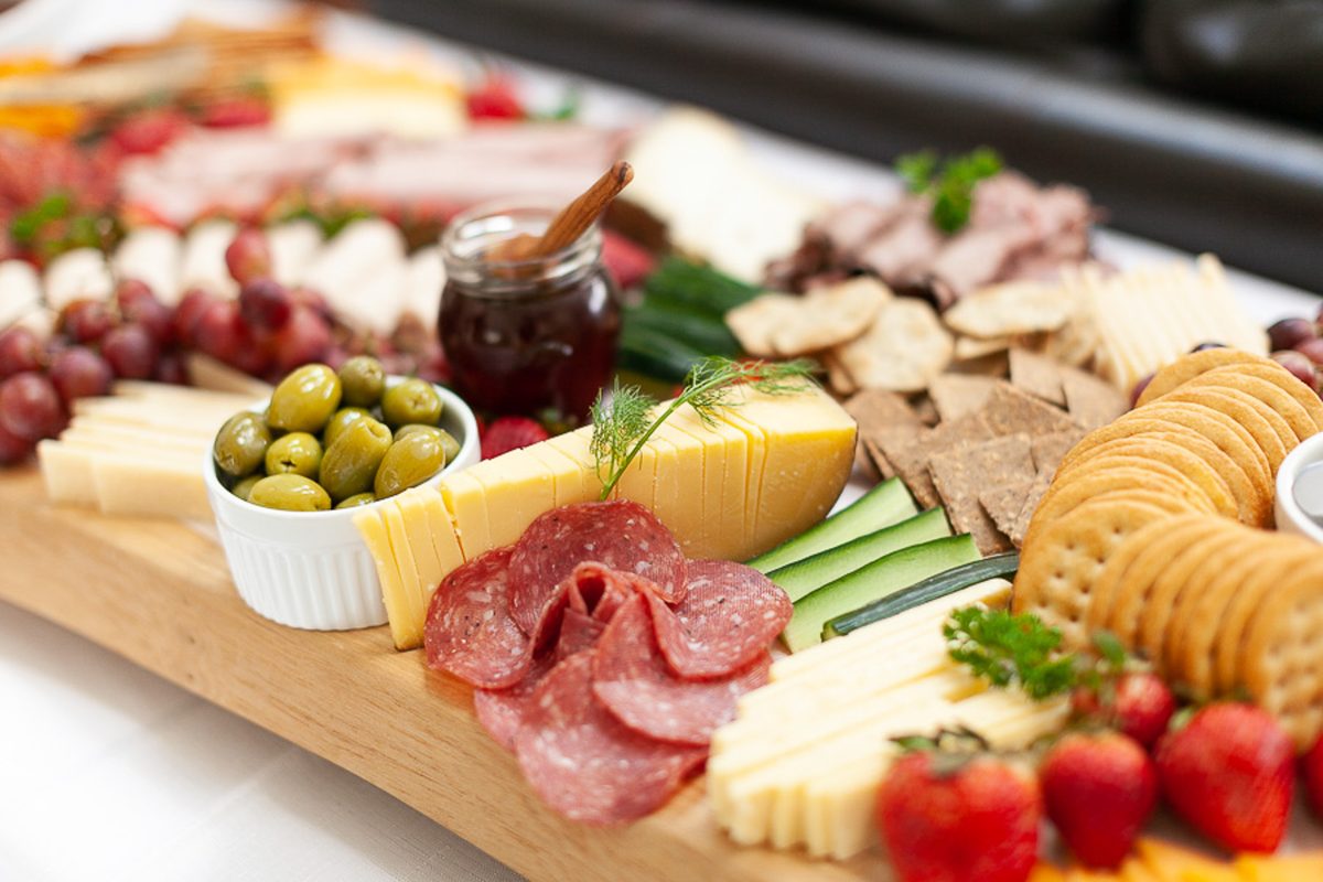 A long wooden charcuterie board loaded with sliced salami, cheeses, crackers, a bowl of green olives, a jar of jam, grapes and str