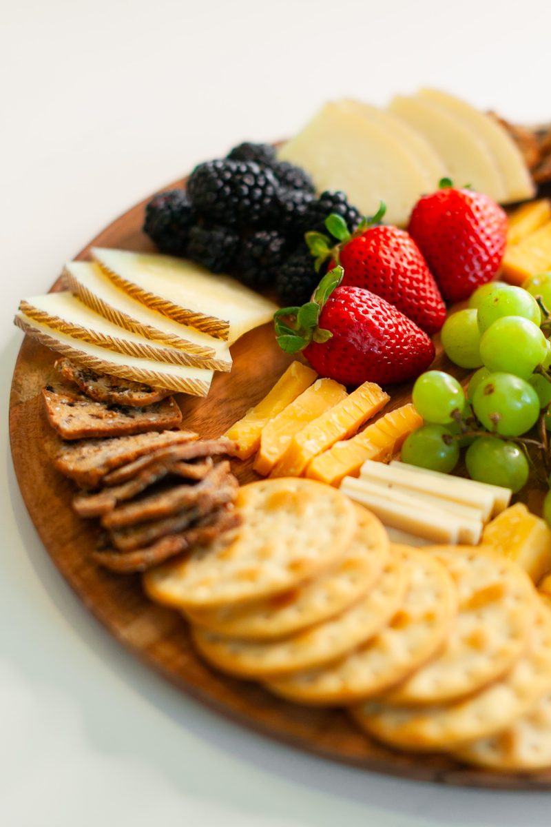 A tight vertical view of a round wooden board arranged with sliced cheeses, cheddar sticks, round and seeded crackers, blackberrie