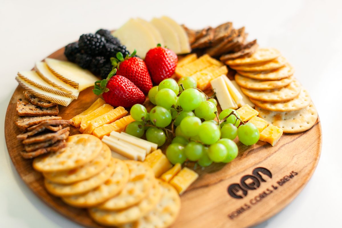 A round wooden board of sliced cheeses, crackers, green grapes, strawberries and blackberries on a white surface, with an engraved