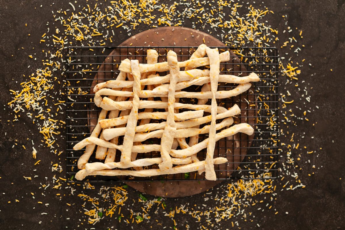 Baked cheese breadsticks woven into a lattice on a round dark wood board set on a cooling rack, with grated cheese and herbs scatt
