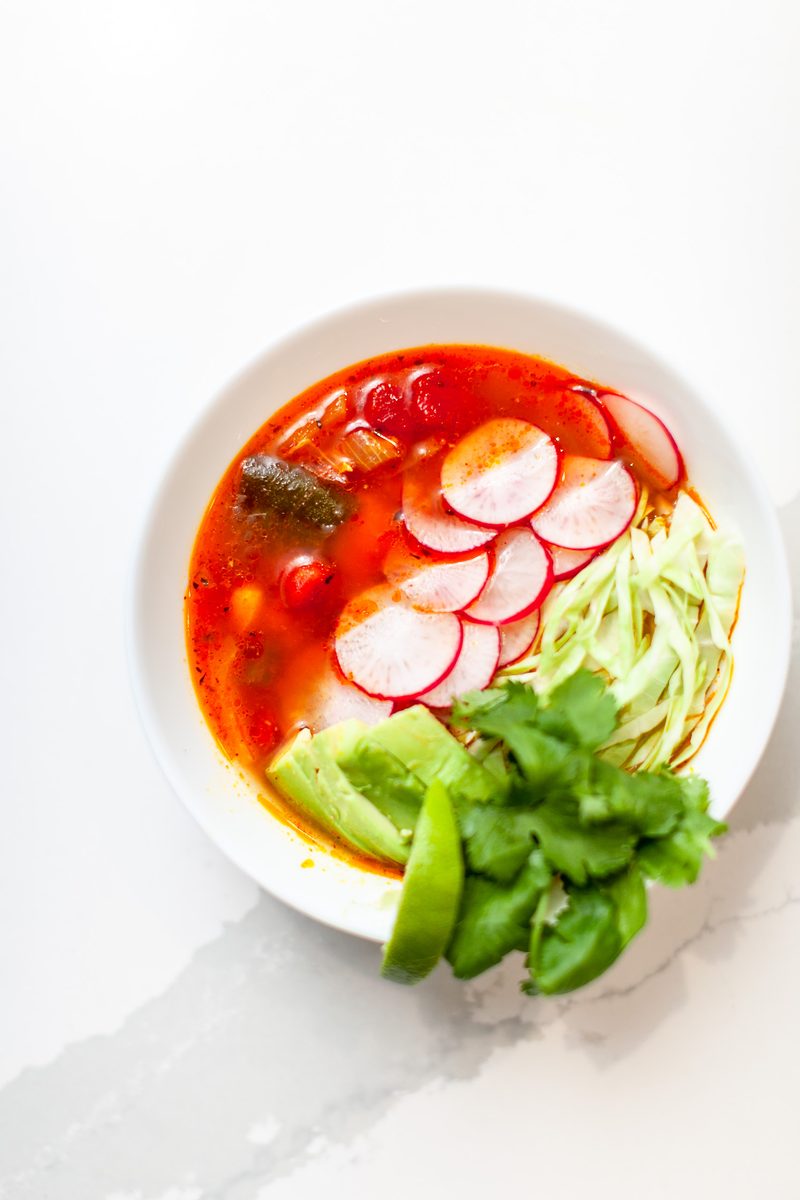 A white bowl of red-broth cold noodles with radish slices, shredded vegetables and green snap peas shot from above on a white marb