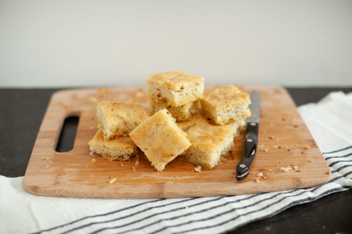 Cut squares of golden cornbread piled on a wooden cutting board with a knife, set on a striped linen towel over a dark counter.
