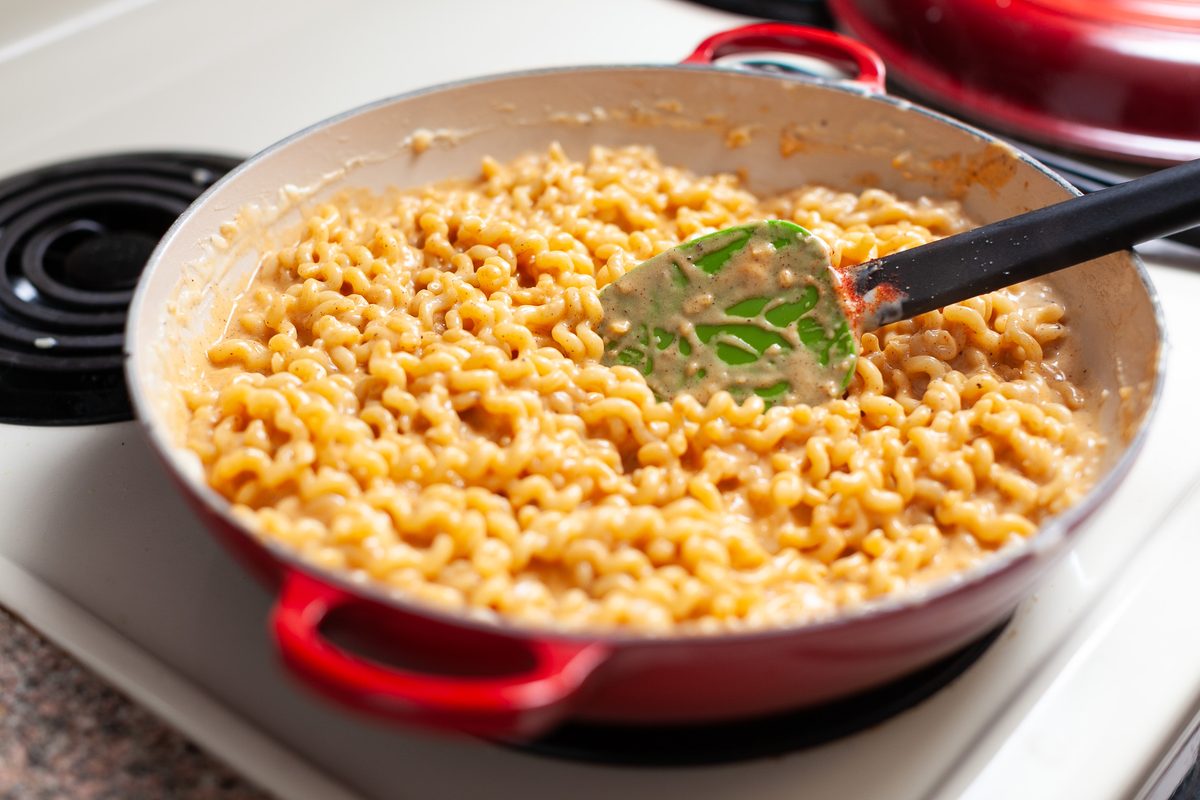 A red enameled skillet of creamy macaroni and cheese with a green silicone spatula resting in it, sitting on a white electric stov
