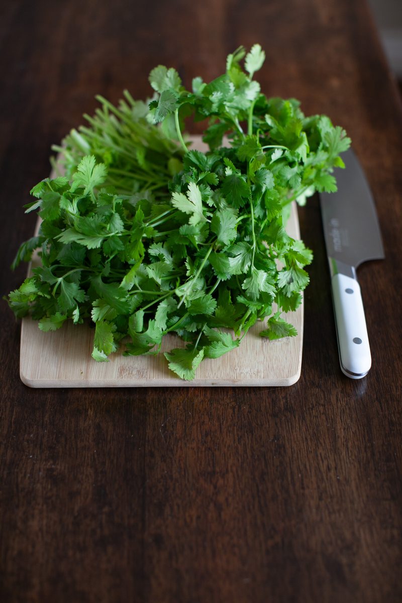 A fresh bunch of cilantro standing on a small wooden cutting board next to a chef's knife on a dark wood table.