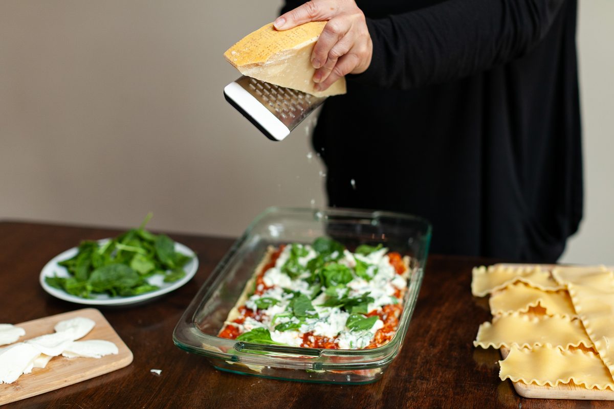 A woman in black grating parmesan over an unbaked lasagna layered with tomato sauce, ricotta and spinach in a glass baking dish, r