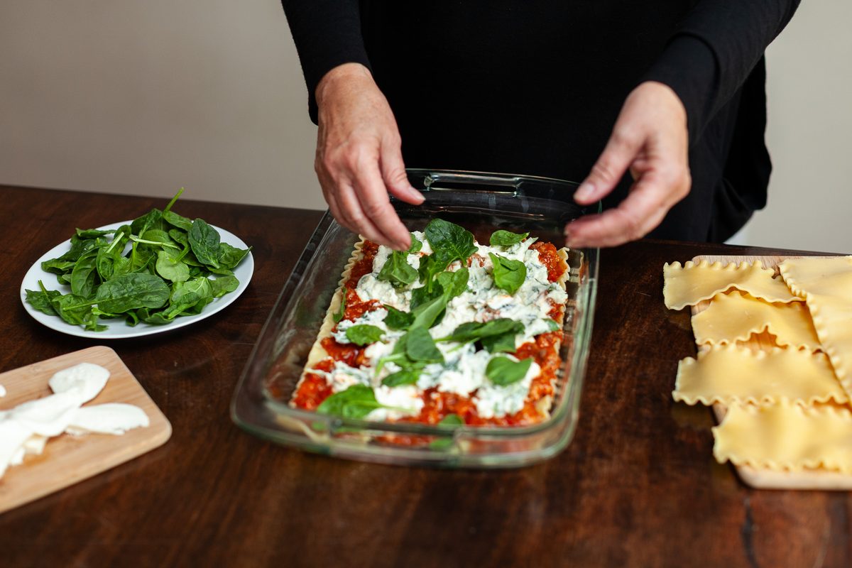Two hands laying fresh spinach leaves over the ricotta and tomato layer of an unbaked lasagna in a glass dish, raw noodles and a p
