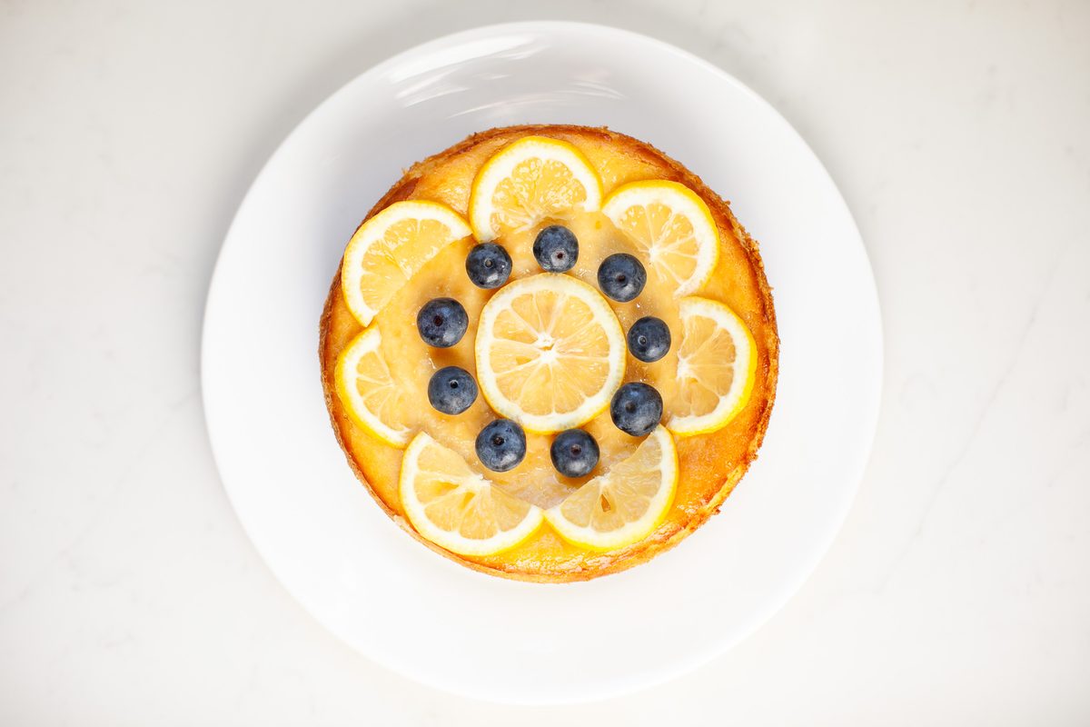 Overhead view of a small round golden lemon ricotta cake on a white plate, ringed with thin lemon slices and topped with a flower 