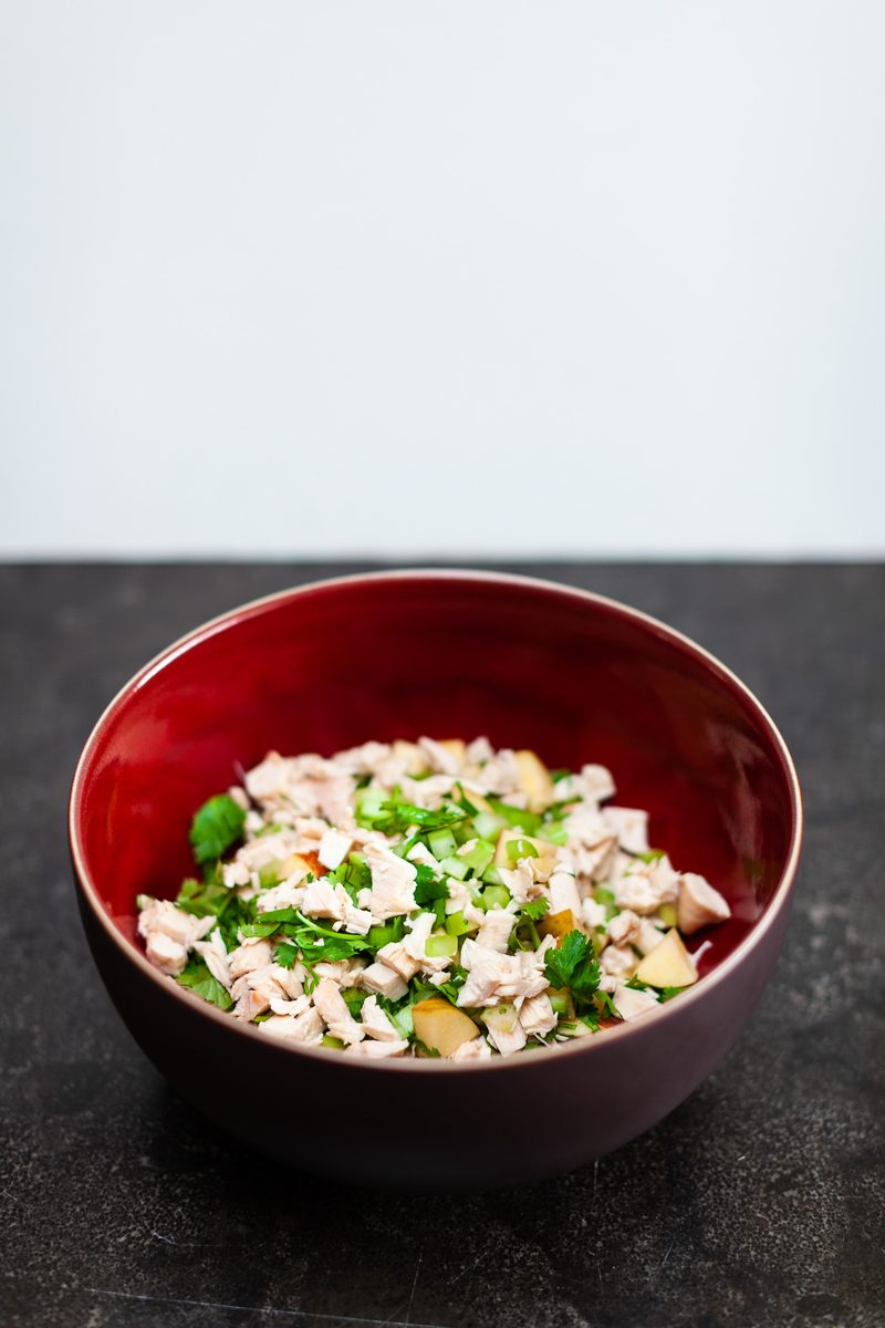 A dark red bowl of chopped chicken salad with diced apple and fresh herbs sitting on a dark slate counter against a pale wall, lot