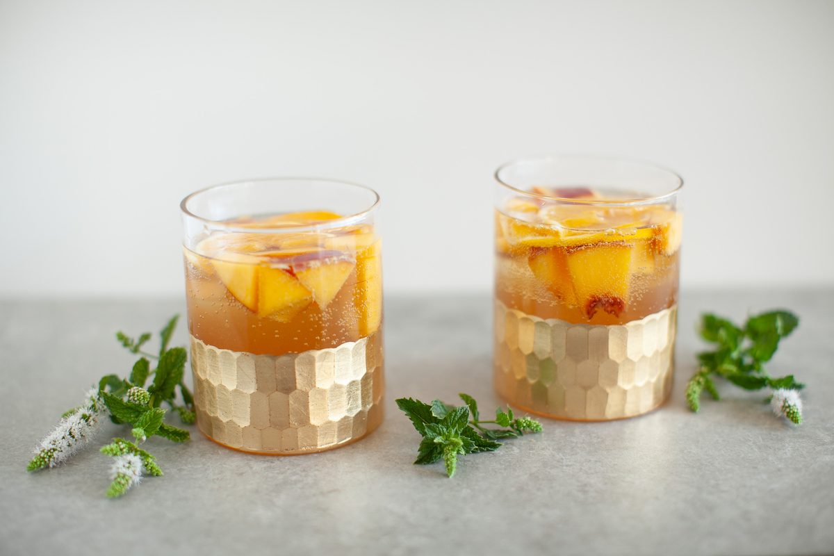 Two short gold-banded tumblers of amber peach and orange sangria with fruit slices inside, on a grey stone surface scattered with 