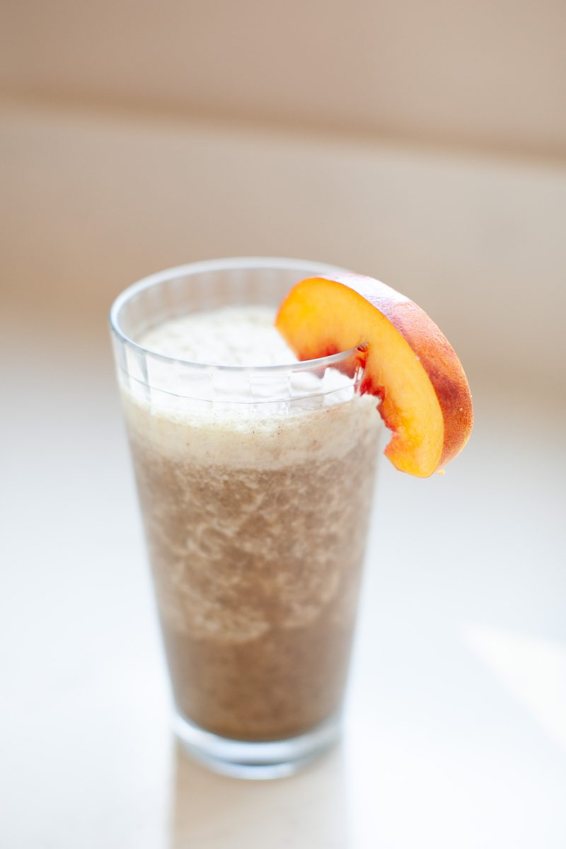 A tall clear glass of thick pale peach smoothie garnished with a fresh peach slice on the rim, on a white surface against a soft b