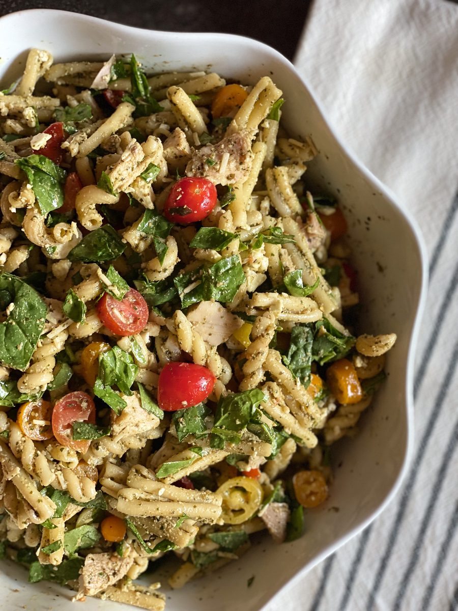 A white scalloped serving bowl of pesto pasta salad with short pasta, halved cherry tomatoes, torn basil and spinach, olives and p