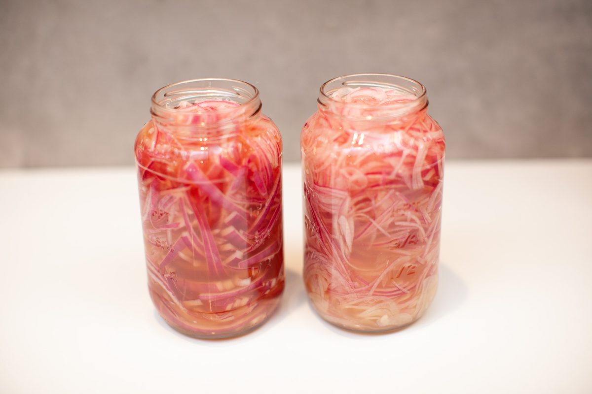 Two glass jars filled with vivid pink pickled red onions on a white counter against a soft grey backdrop, shot straight on.