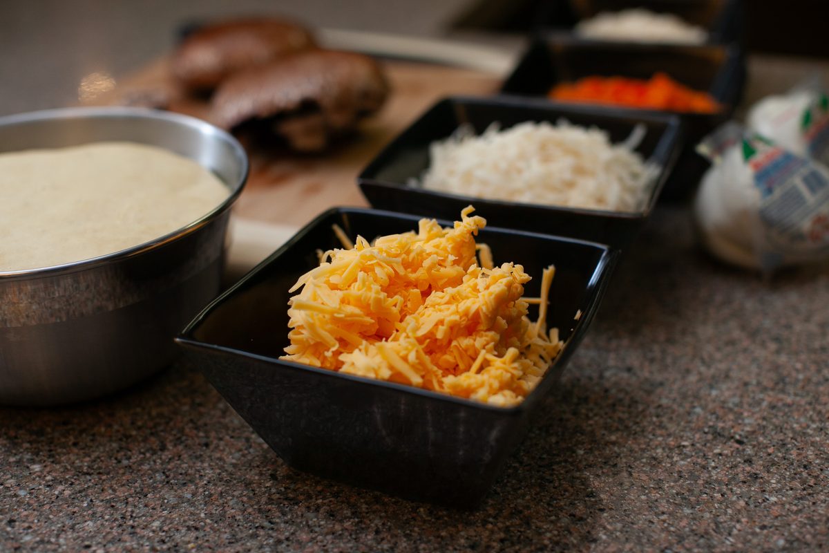 Pizza prep on a granite counter - black square bowls of shredded cheddar and shredded mozzarella, a metal bowl of dough or batter,