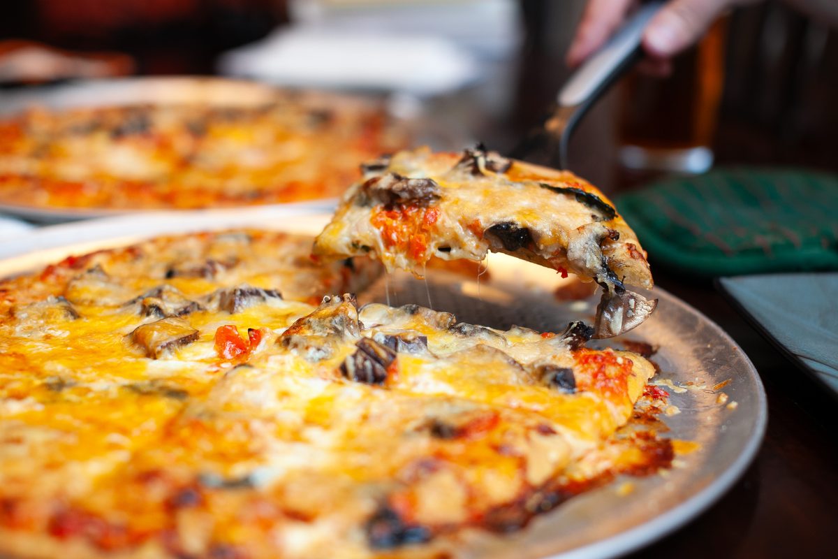 A hand lifting a cheesy slice from a thin-crust pizza topped with mushrooms, olives and tomato on a metal pizza pan, second pizza