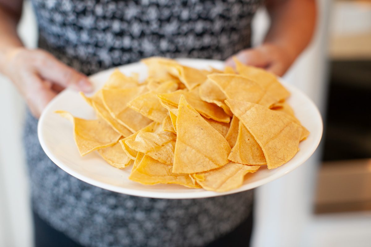 A person in a patterned dark top (head out of frame) holding out a white plate piled with pale homemade corn tortilla chips in a k