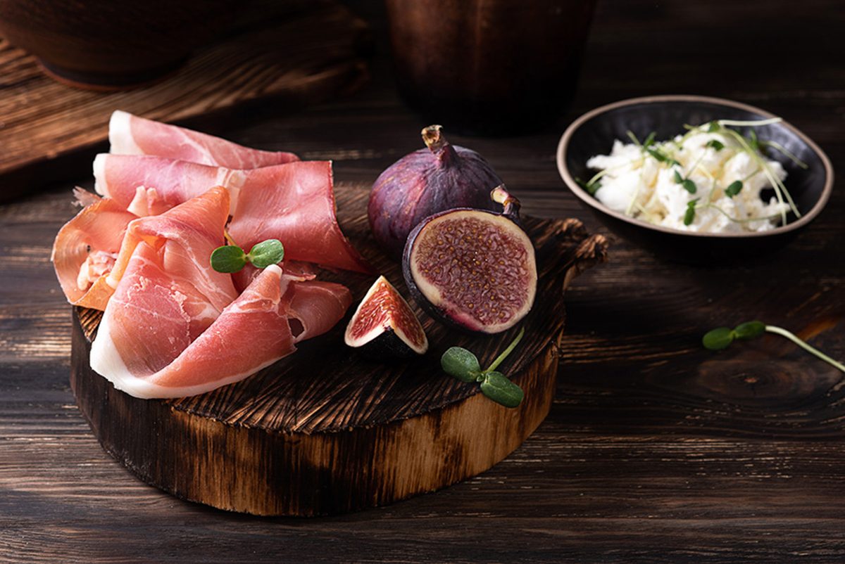 A dark round wooden board with folded slices of prosciutto, halved fresh figs and mint leaves, next to a dark bowl of herbed soft 