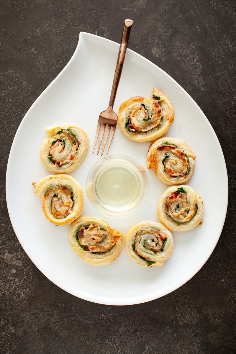 Overhead shot of prosciutto and spinach puff pastry pinwheels on a white teardrop-shaped platter with a small well of dip, on a da