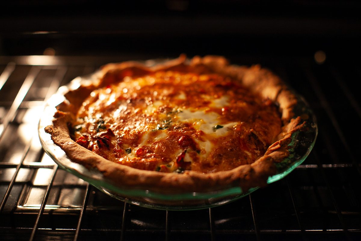 A quiche with a browned crimped crust and bubbling cheesy top baking on the wire rack inside a dark oven, shot through the open do