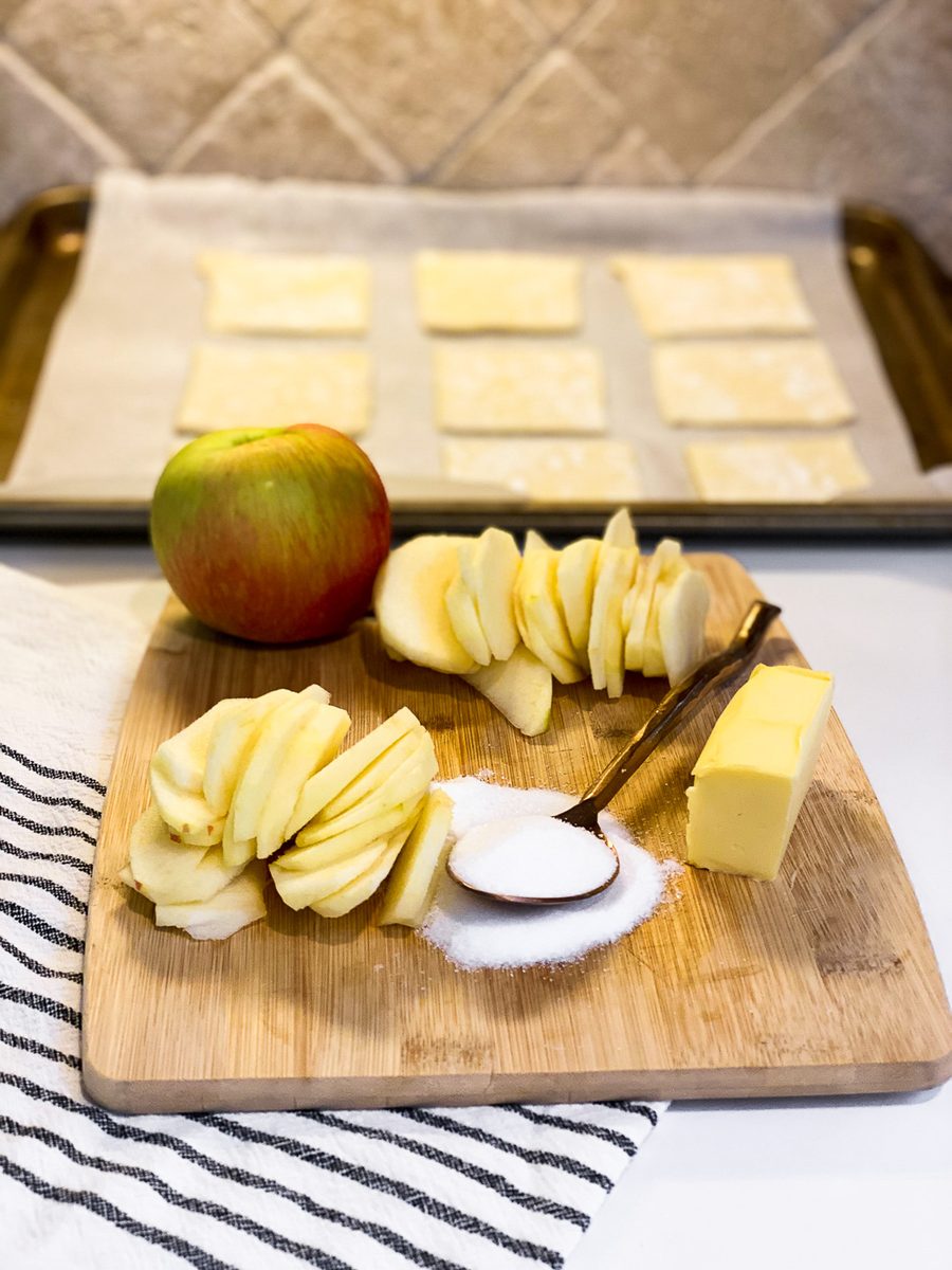 A wooden board with two fanned piles of thinly sliced apple, a whole apple, a block of butter, a spoon of sugar and a vanilla bean