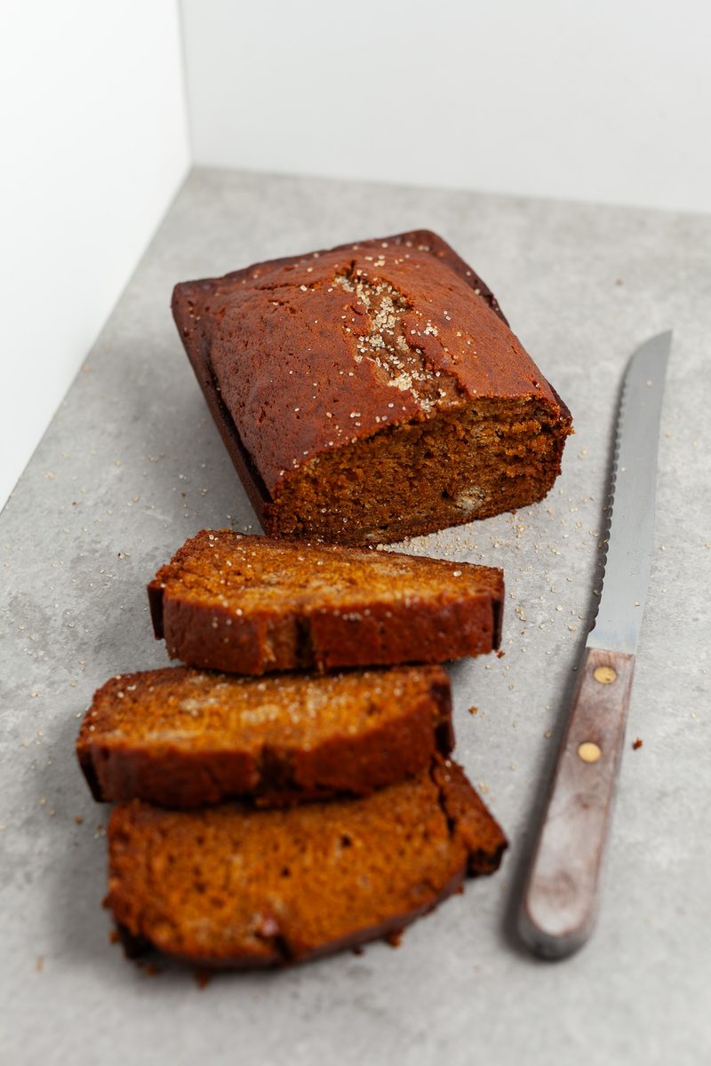 A dark spiced pumpkin bread loaf with three thick slices fanned in front and a wood-handled bread knife alongside, on a pale grey