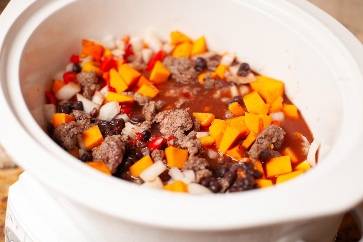 Close overhead view into a white slow cooker crock filled with uncooked chili - browned ground beef, cubed butternut squash, black