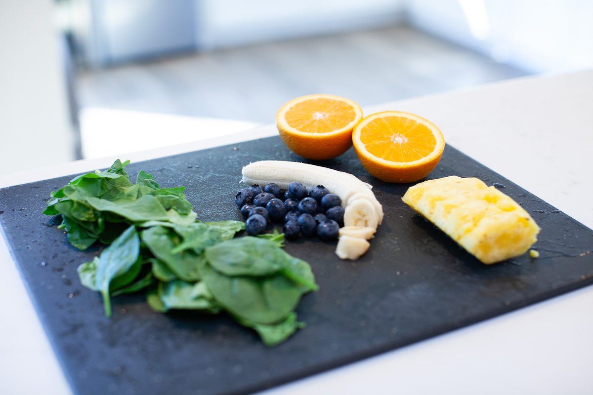 A dark slate board on a counter arranged with fresh spinach leaves, blueberries, banana pieces, chopped mango and two orange halve