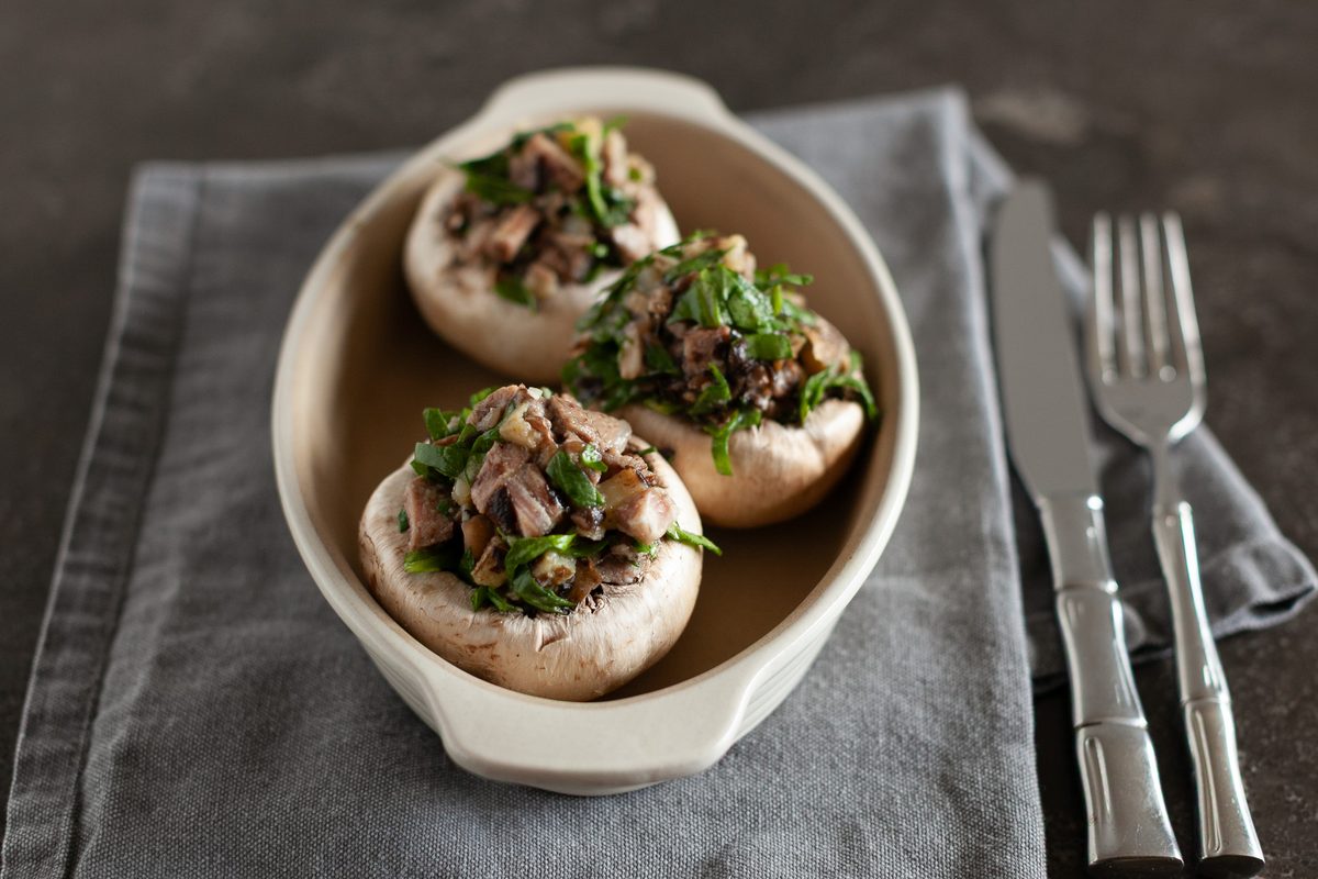 Close view of three plump white mushroom caps filled with chopped mushroom stems, parsley and nuts in a cream oval baker on a grey