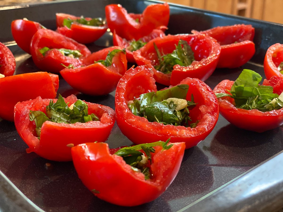 A dark metal baking tray crowded with halved and quartered red bell peppers filled with chopped fresh herbs, basil and garlic, unr