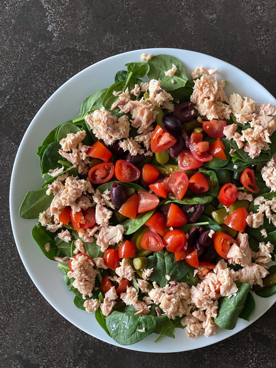 Overhead shot of a salad of flaked tuna, baby spinach leaves, chopped tomatoes and dark olives in a shallow white bowl on a dark g