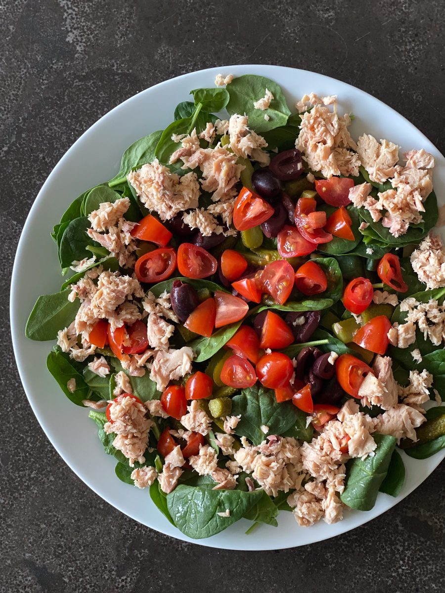 Overhead shot of the same flaked tuna, spinach, chopped tomato and olive salad in a white bowl on a dark slate background, slightl