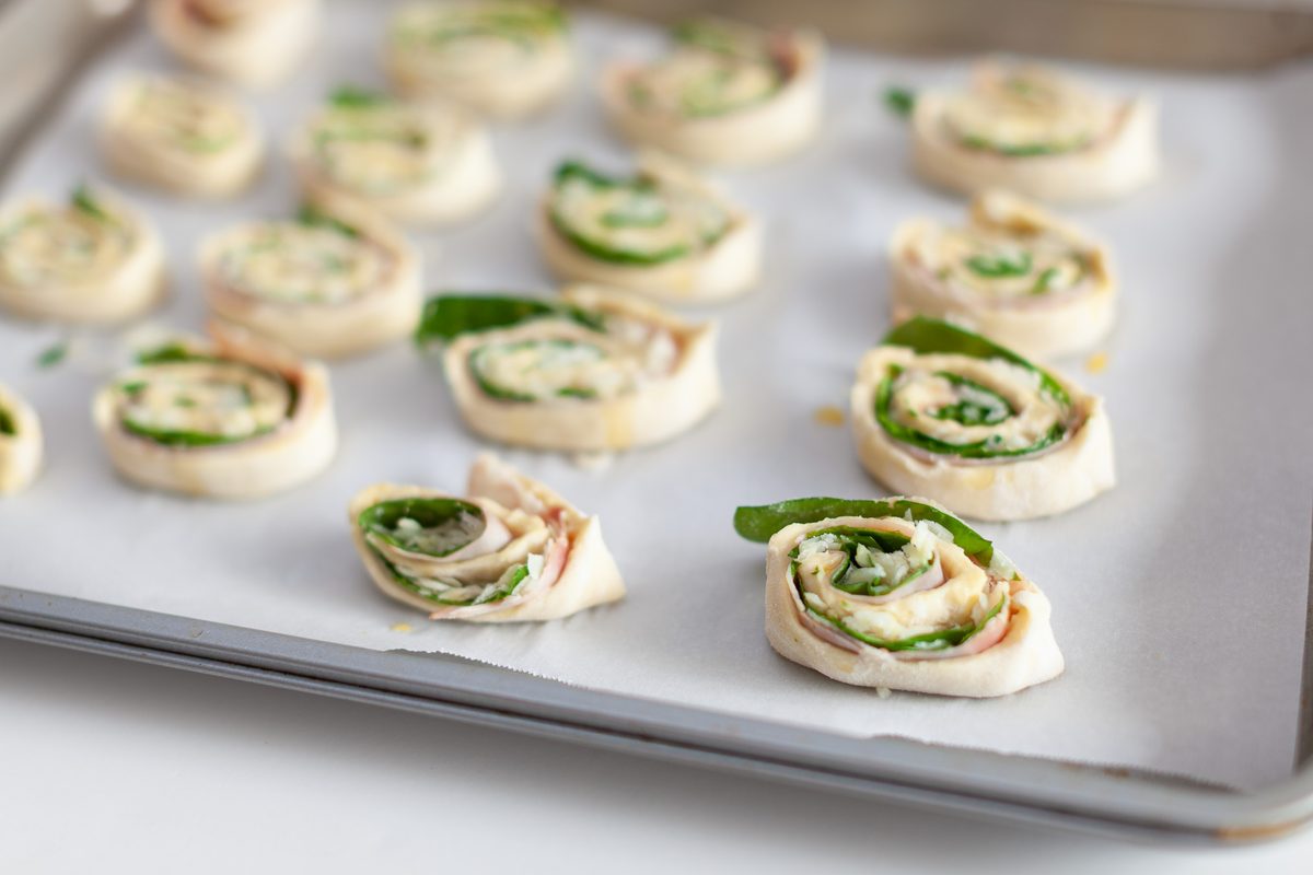 Rows of assembled tortilla pinwheels filled with deli turkey, cream cheese and spinach laid out on a parchment-lined sheet pan, no
